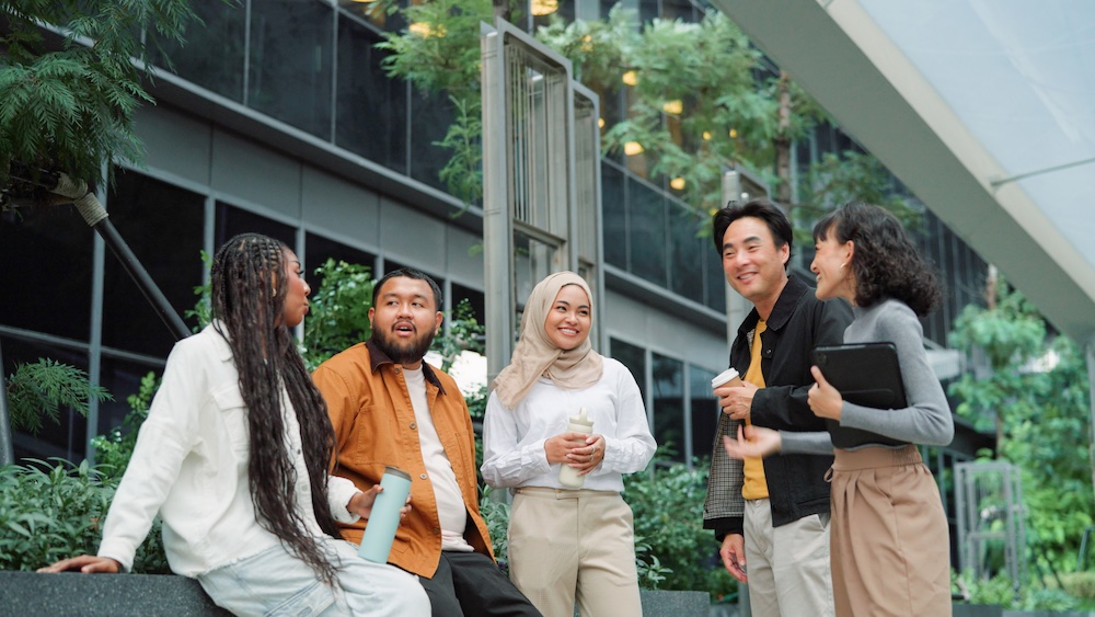 Five people, diverse in appearance and dress, stand and sit together outdoors by a modern building, smiling and talking. Some hold drinks and a tablet, surrounded by greenery and contemporary architecture.