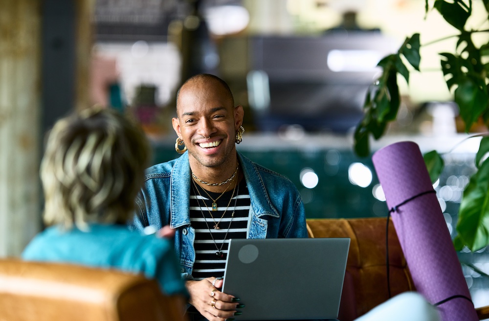 A person with short hair and multiple earrings smiles while holding a laptop and talking to another person in a casual, indoor setting with plants and a yoga mat nearby.