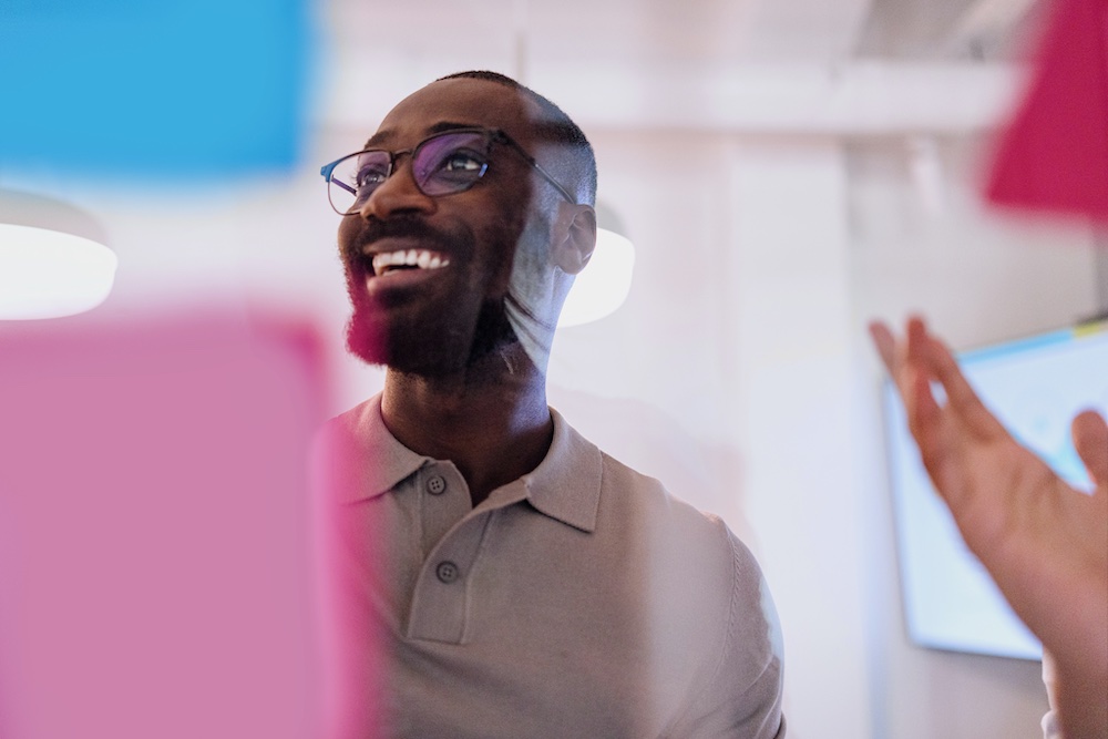 A man wearing glasses and a gray polo shirt smiles while standing behind colorful sticky notes in an office setting. His expression appears enthusiastic and engaged.