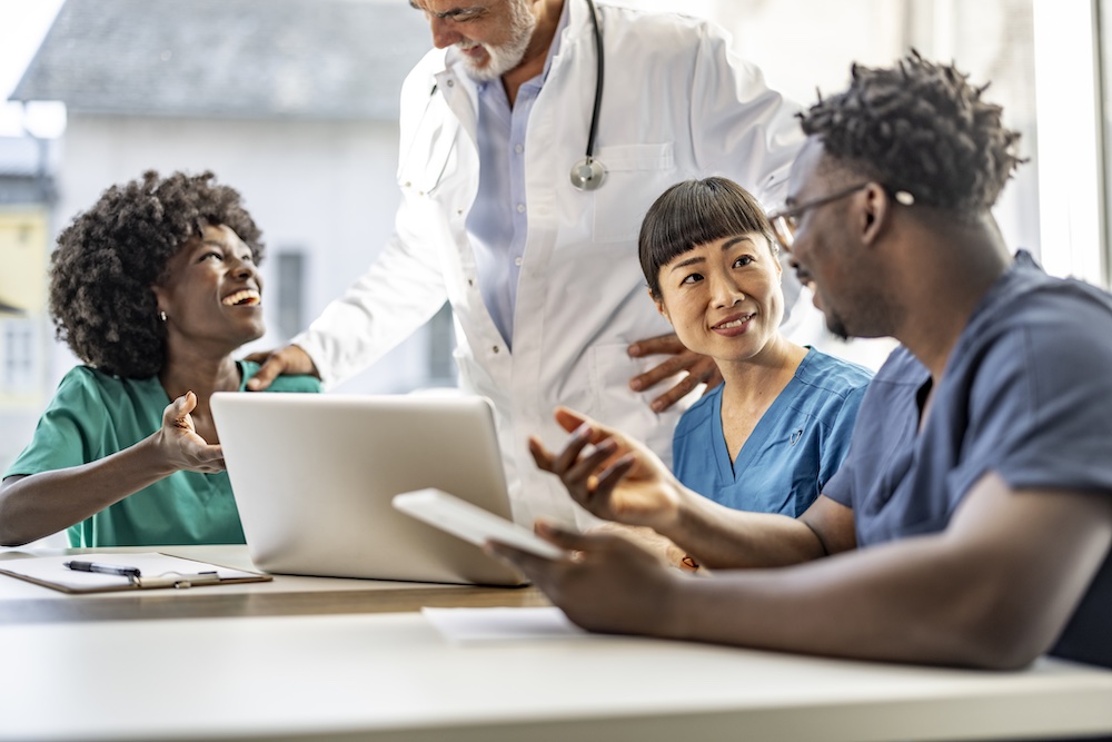 Four medical professionals, including a doctor in a white coat and three nurses in scrubs, sit around a table with a laptop and tablet, smiling and having a discussion in a bright room.