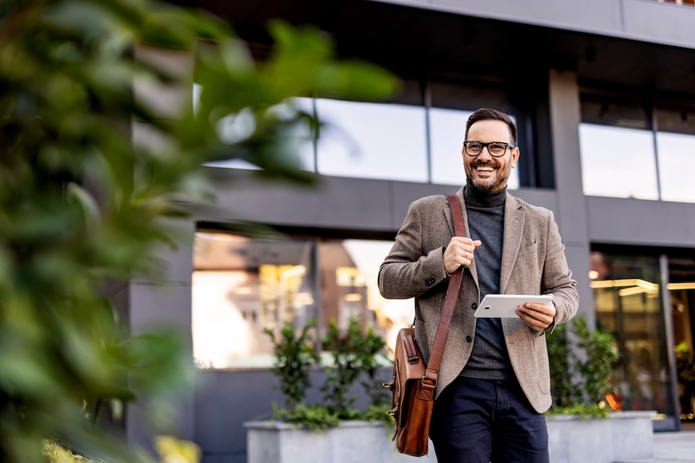 A smiling man with glasses and a beard, wearing a brown blazer and carrying a shoulder bag, stands outside a modern building holding a tablet. Green plants are in the foreground.