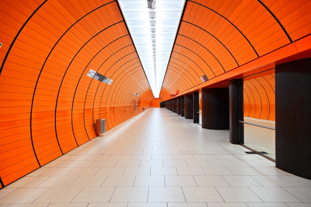 A modern, empty underground subway tunnel with bright orange curved walls, a white tiled floor, and black columns, creating a symmetrical, futuristic appearance. Signs and lights line the ceiling.