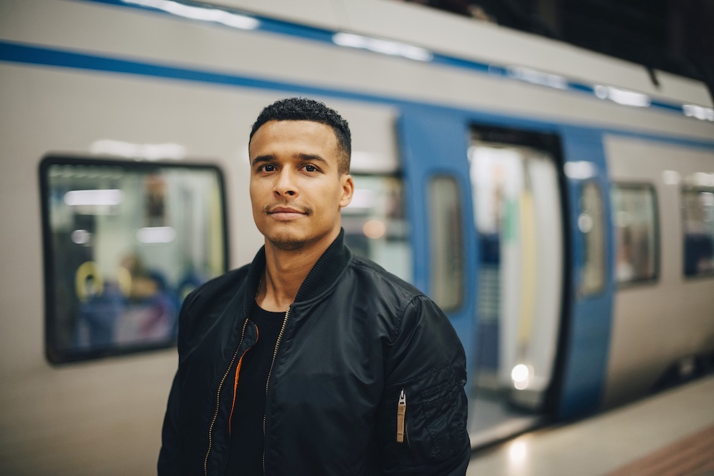 A man wearing a black jacket stands on a train platform, looking at the camera, with a blue and white train stopped behind him and its doors open.