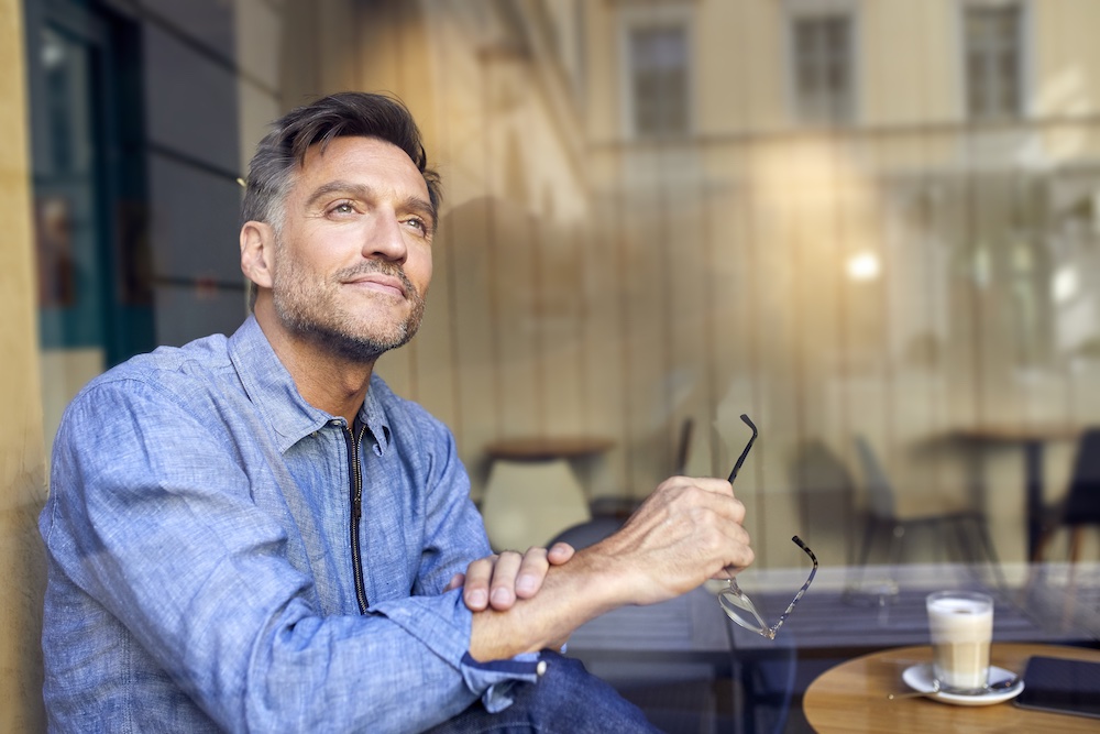 A middle-aged man in a blue shirt sits at a café table by a window, holding glasses in one hand and looking thoughtfully outside. A cup of coffee is on the table beside him.