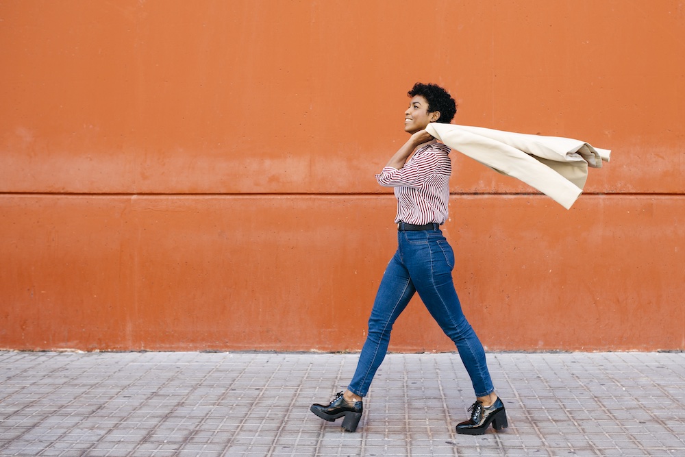 A woman in jeans and heeled boots walks confidently along a tiled sidewalk, smiling and holding a beige coat over her shoulders, with an orange wall in the background.