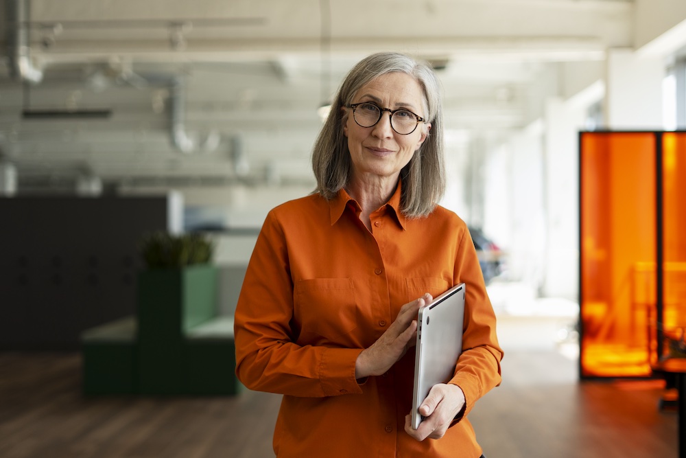 An older woman with long gray hair and glasses, wearing an orange shirt, stands in a modern office holding a tablet and smiling slightly at the camera.