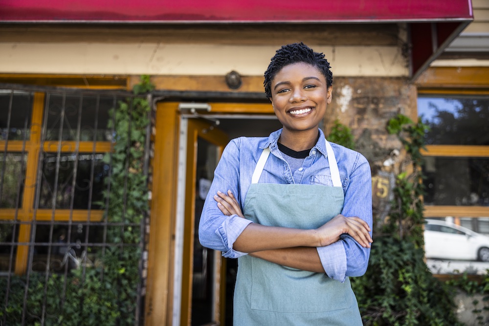 A smiling person wearing a light blue shirt and green apron stands with arms crossed in front of a building with large windows and a red awning.