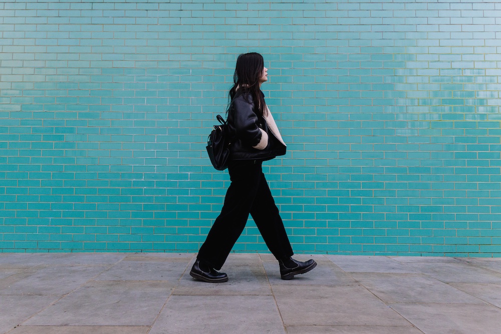 A person with long dark hair, dressed in black and carrying a black backpack, walks past a turquoise brick wall on a paved sidewalk.