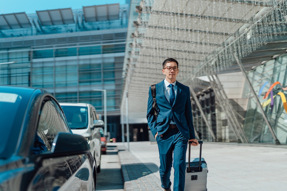 A man in a blue suit walks outside a modern building, pulling a suitcase with one hand and carrying a backpack. There are cars parked alongside the walkway. The weather appears sunny and clear.