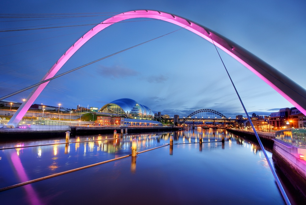 A modern, illuminated arched bridge glows with pink lights over a calm river at dusk, reflecting city buildings, lights, and a domed glass structure along the waterfront.