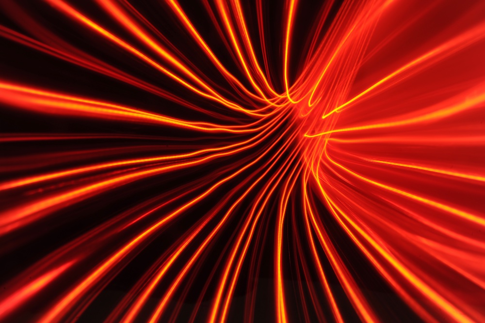 Red and orange light trails curve and stretch across a black background, creating a dynamic, abstract pattern that resembles motion or energy flowing toward a central point.