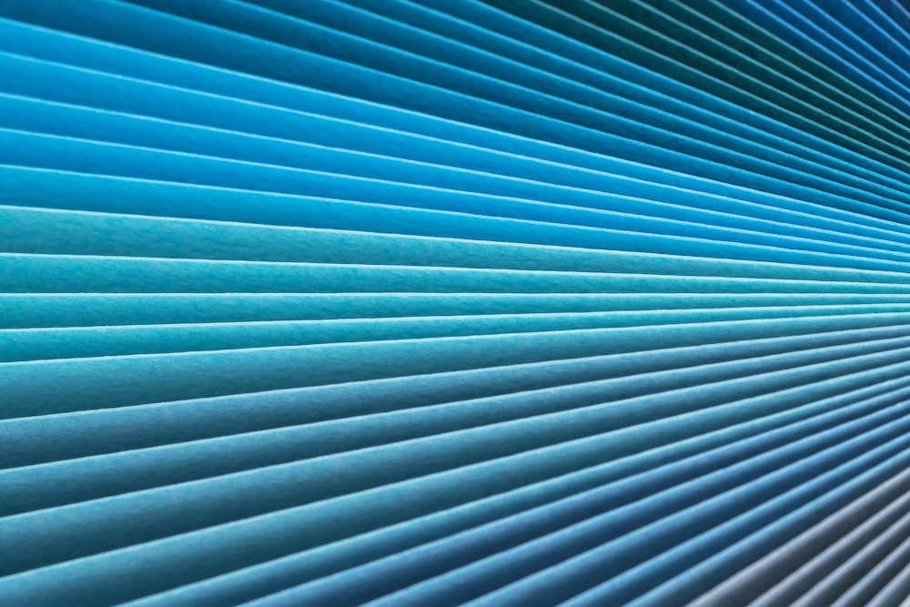 Close-up of evenly spaced, diagonal blue paper sheets or slats creating a geometric, abstract pattern with varying shades of blue and teal, fading from light to dark.