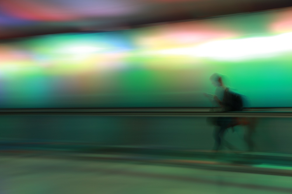 A blurred silhouette of a person walking with a backpack and looking at a phone, set against a vibrant, multicolored, and softly glowing background.