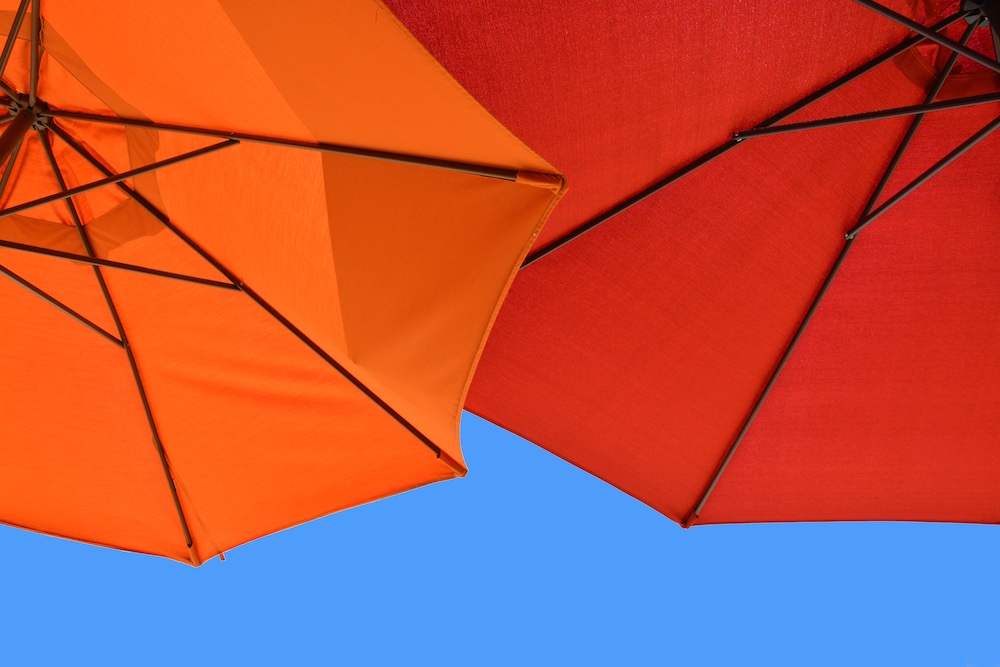 Two open umbrellas, one orange and one red, are seen from below against a clear blue sky. The umbrella frames and fabric panels are visible in the image.