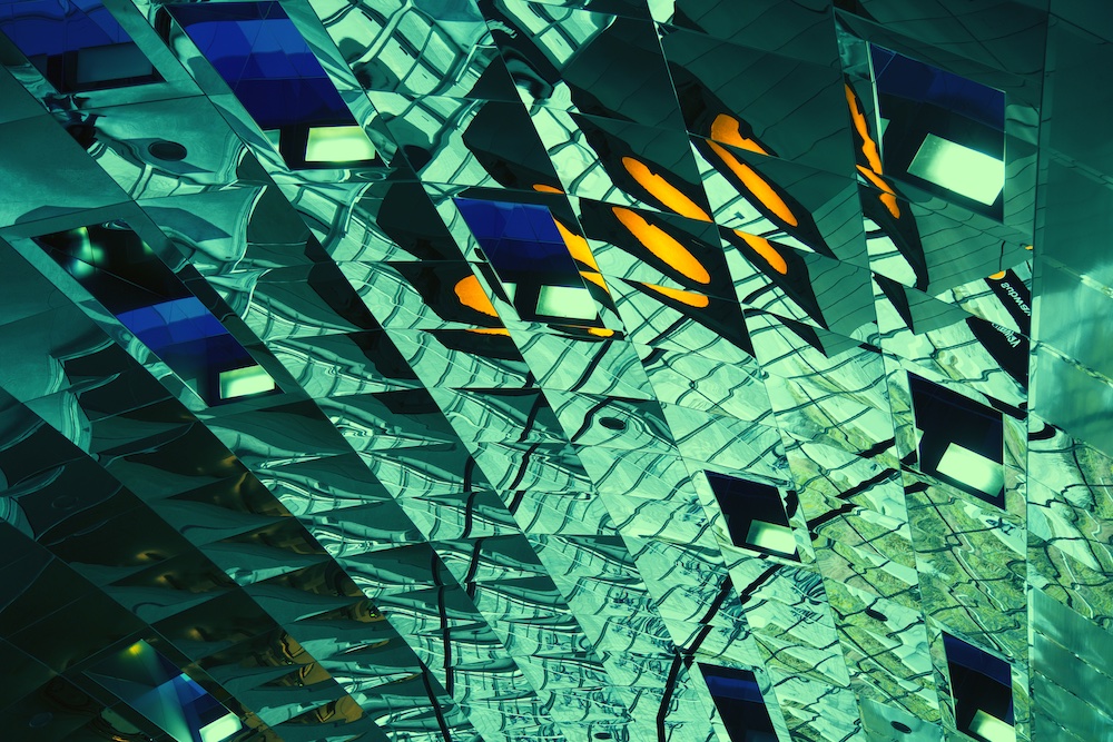 Abstract image showing a pattern of green and blue mirrored panels with geometric shapes and reflections, interspersed with bright yellow-orange accents, creating a kaleidoscopic, futuristic appearance.