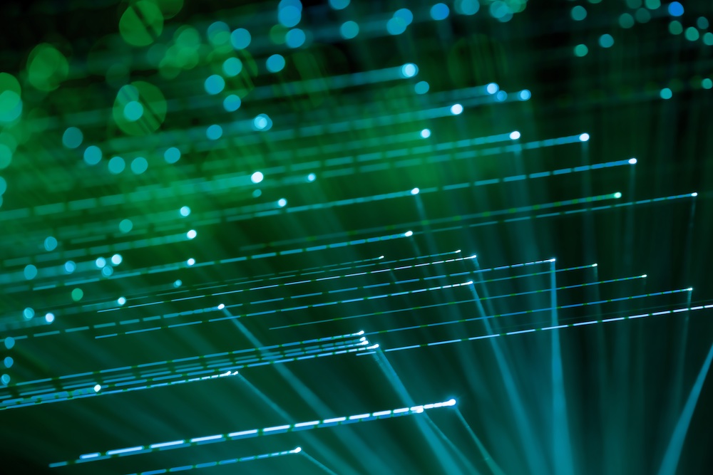 Green and blue fiber optic cables glowing in the dark, creating a dynamic pattern of light streaks and dots against a blurred background.