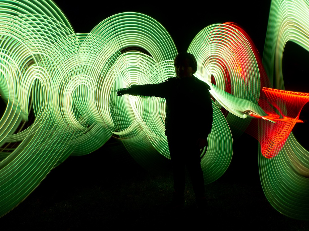 A silhouette of a child stands with an outstretched arm against a background of swirling green and red light trails in the darkness.