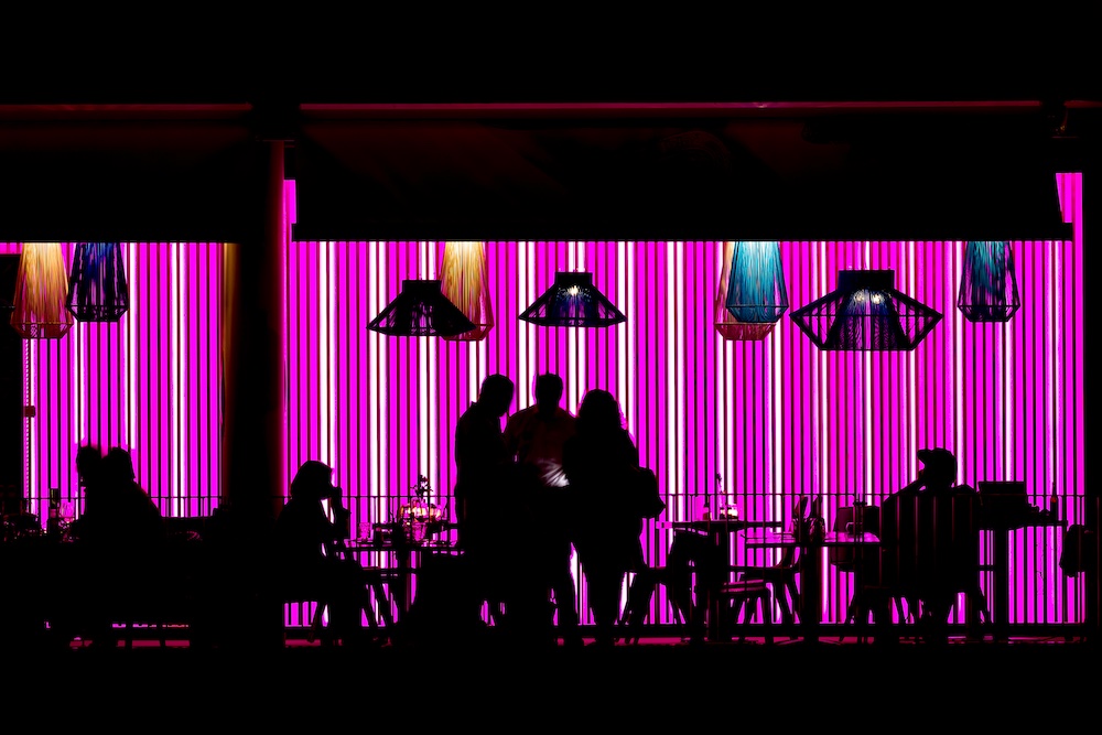 Silhouettes of people sitting and standing at tables in a restaurant or café, with a vibrant pink striped neon light wall and hanging lamps in the background.