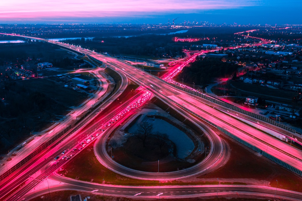 Aerial view of a busy highway interchange at dusk, with red and white vehicle lights forming glowing trails as cars move along multiple intersecting roads, surrounded by fields, houses, and distant city lights.
