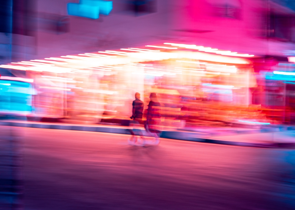 Two blurred figures walk past a brightly lit building at night, surrounded by vibrant neon pink, blue, and orange lights, creating an abstract, motion-filled city scene.