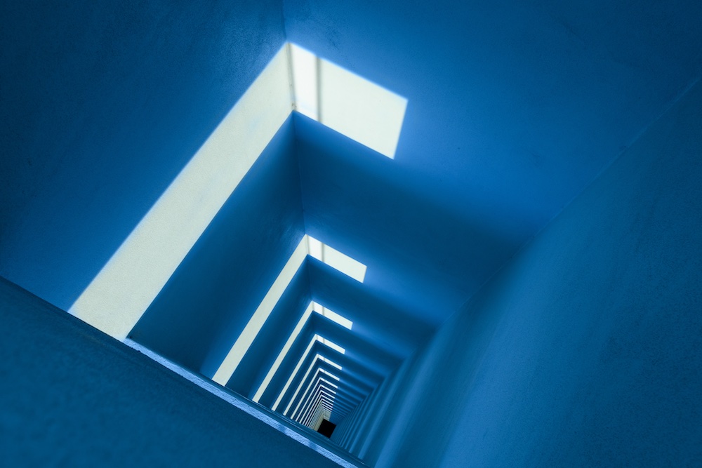 A tilted view of a blue corridor with rectangular skylights casting bright geometric patterns of light and shadow along the walls and floor, creating an abstract, tunnel-like effect.