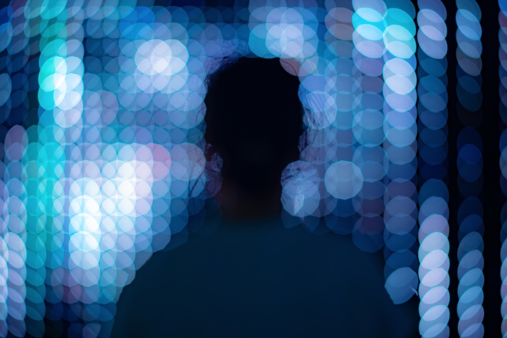 A silhouetted figure stands facing a background filled with blurry blue and white bokeh lights, creating a dreamy, abstract atmosphere.