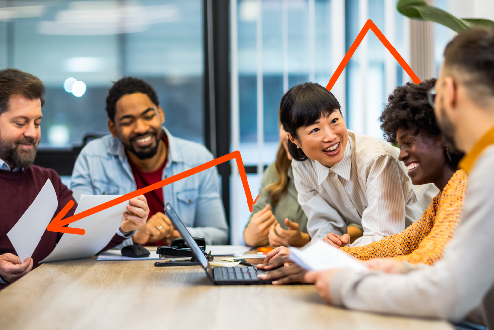 A diverse group of colleagues sit around a table, smiling and discussing papers and a laptop, with an orange arrow drawn above them indicating teamwork and progress.