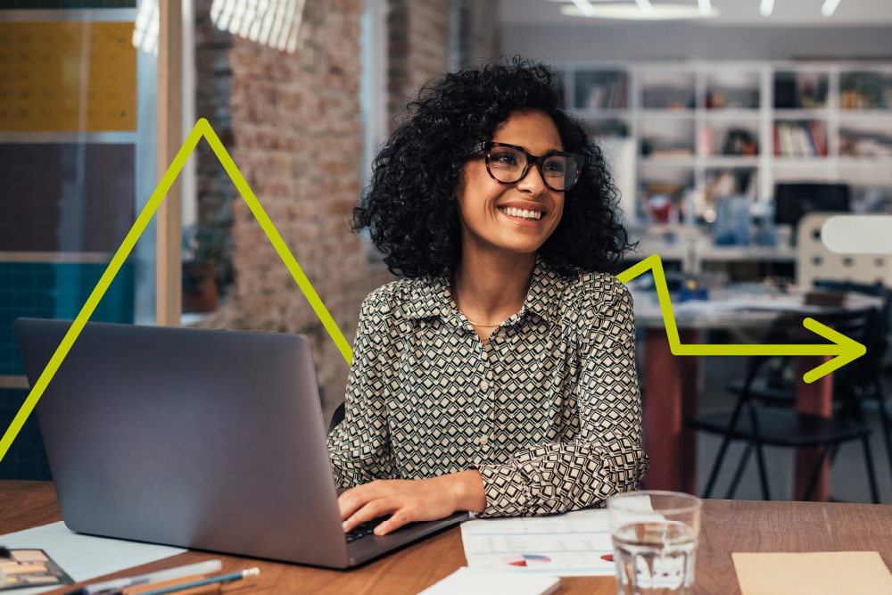 A woman with curly hair and glasses smiles while working on a laptop at a desk with papers and a glass of water. A yellow arrow and line graph are digitally drawn in the background, suggesting progress.