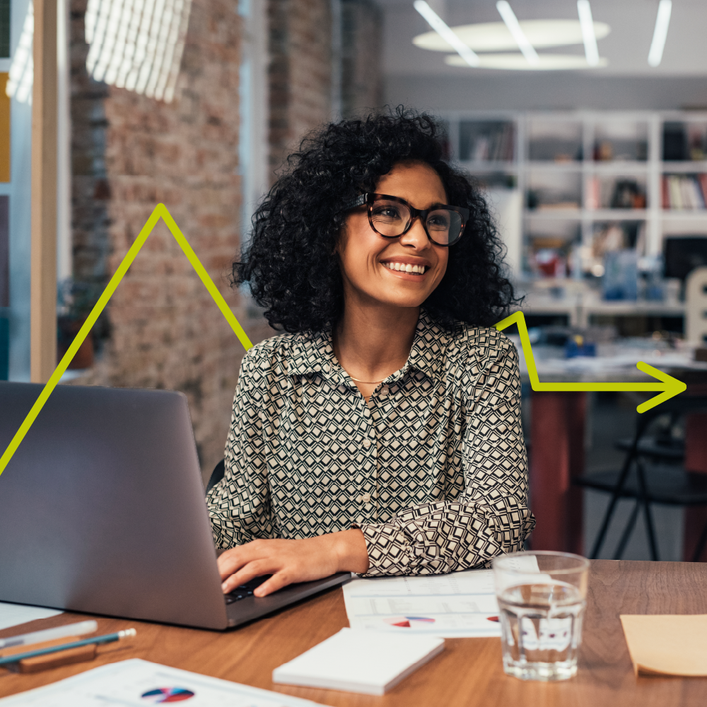 A woman with curly hair and glasses smiles while working on a laptop at a desk in a modern office. A yellow arrow graphic is overlaid, suggesting growth or progress. Papers and a glass of water are on the table.