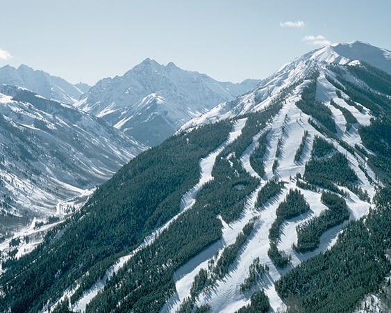 A snow-covered mountain with ski trails winding through pine trees, set against a backdrop of rugged, snow-capped peaks under a clear sky.