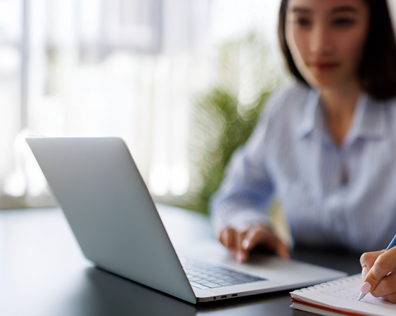 A woman sits at a desk working on a laptop, with one hand on the keyboard and the other writing in a notebook. The background is softly blurred, suggesting a bright indoor setting.