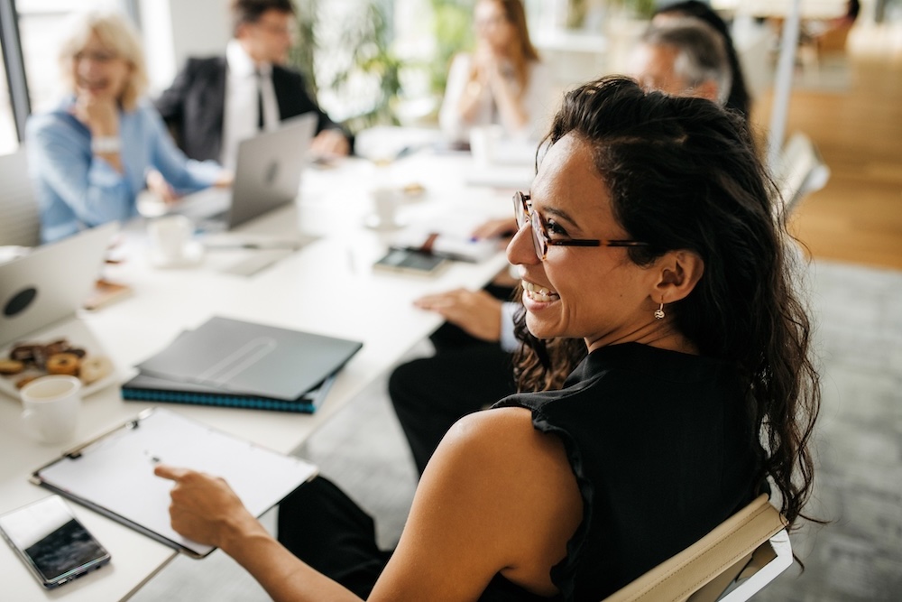 A woman in glasses smiles while holding a notepad at a conference table with colleagues, laptops, and documents during a business meeting.